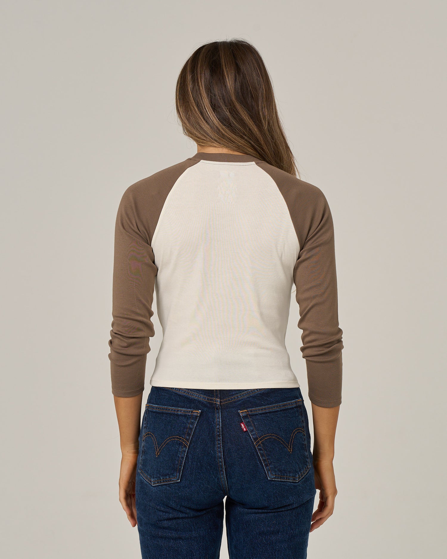 A woman with long brown hair, wearing the Salty Crew Pretty Palms Long Sleeve in Washed Brown and blue jeans, is standing with her back to the camera against a plain background.