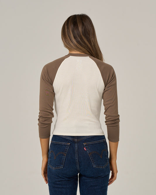 A woman with long brown hair, wearing the Salty Crew Pretty Palms Long Sleeve in Washed Brown and blue jeans, is standing with her back to the camera against a plain background.