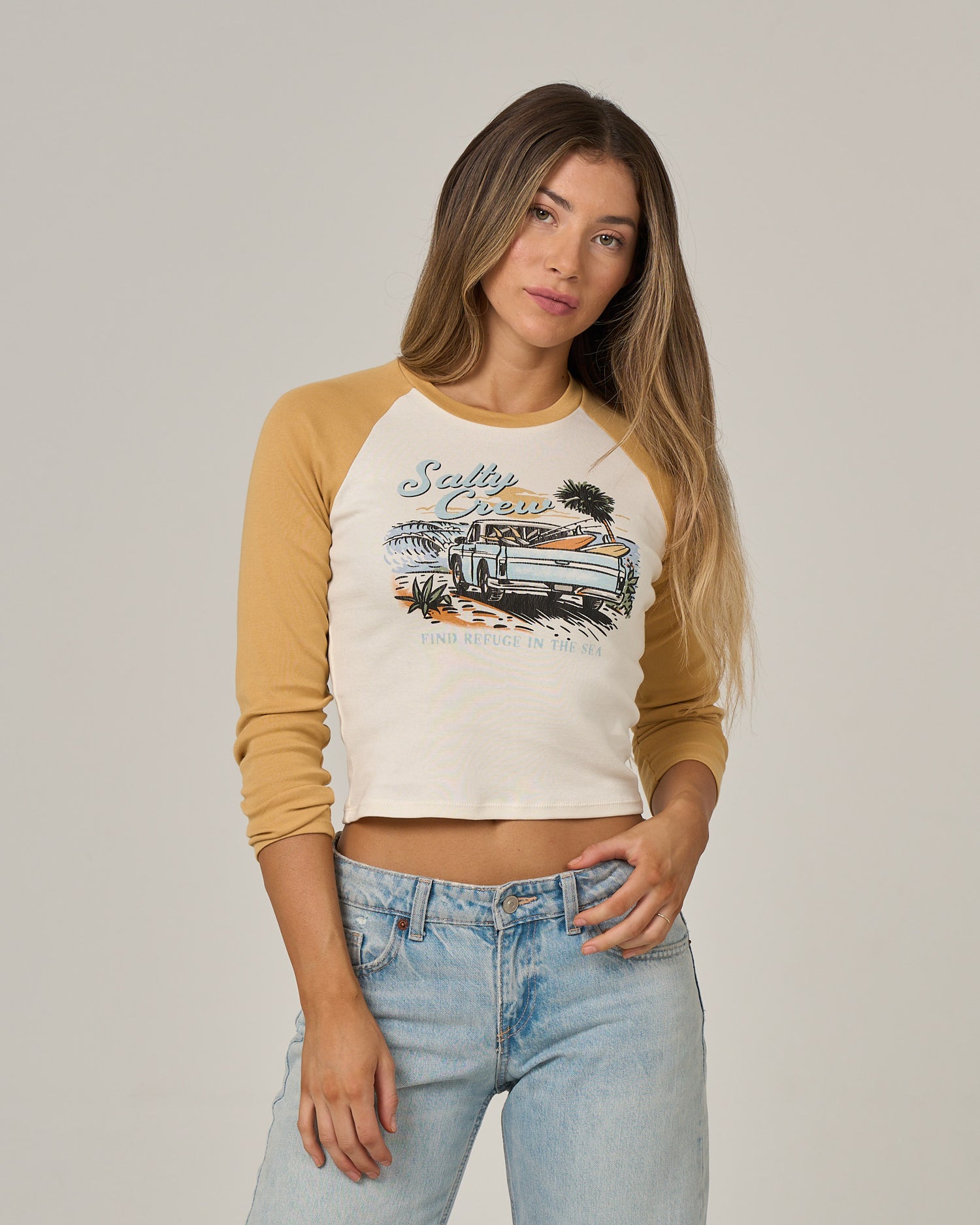 A woman with long hair wears the Salty Crew Truck Bed Long Sleeve - Gold, a cream and tan cotton ribbed tee featuring a screen print of a car and palm trees, paired with light blue jeans against a plain background.