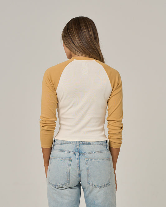 A person with long brown hair, standing facing away against a plain background, is wearing the Salty Crew Truck Bed Long Sleeve - Gold and light blue jeans.