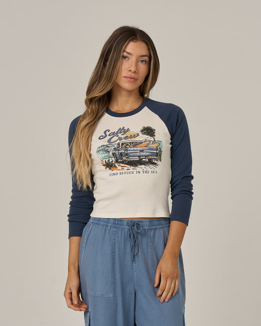 A woman with long hair wears the Salty Crew Truck Bed Long Sleeve shirt in True Navy, which features a beach graphic screen print, paired with blue drawstring pants, and stands against a plain light background.