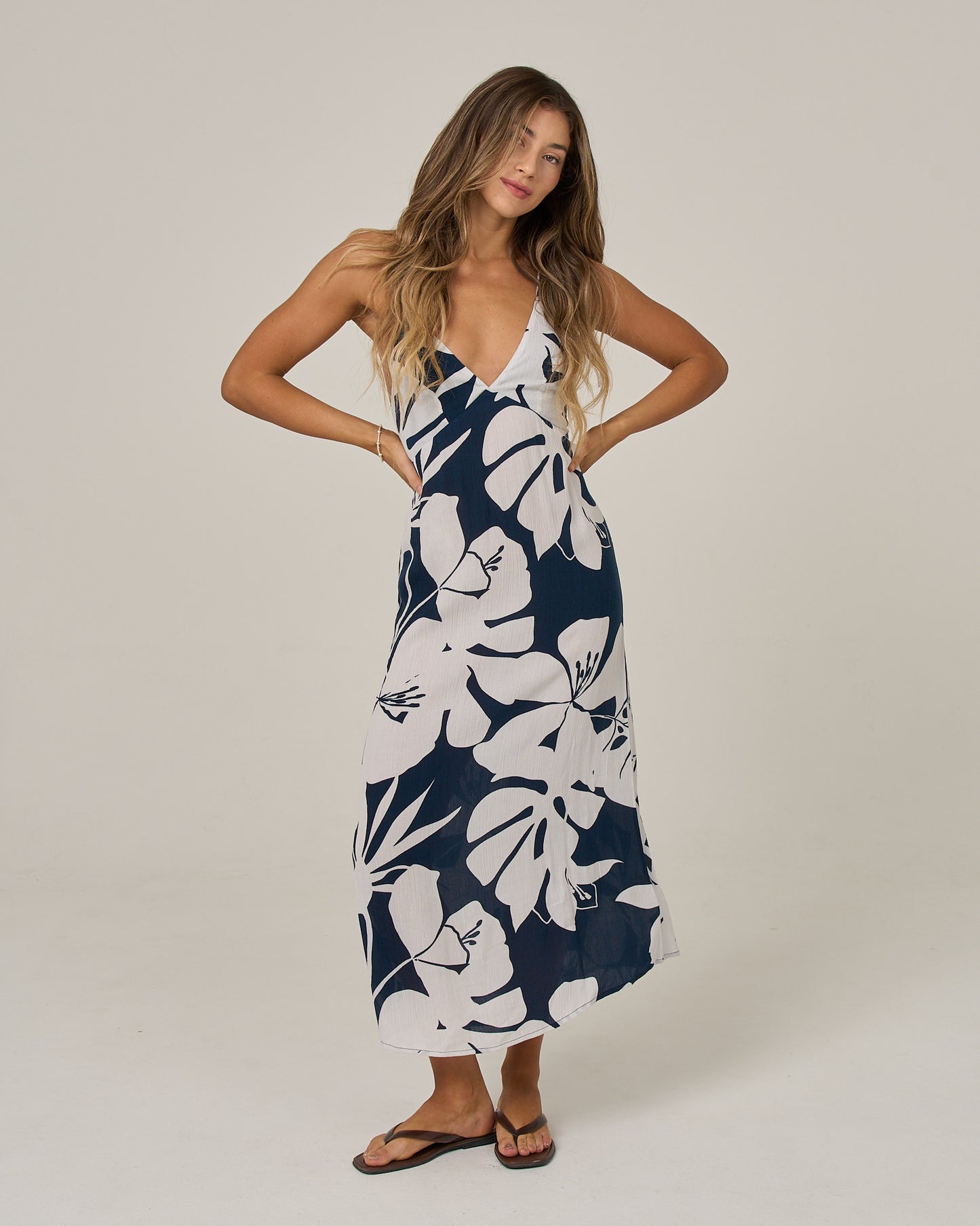 A woman with long wavy hair stands with her hands on her hips against a plain light background, wearing the Salty Crew Graphic Bloom Dress in Faded Mauve—a sleeveless slip dress featuring a deep V-neck, navy and white floral print, cross back straps, and black sandals.