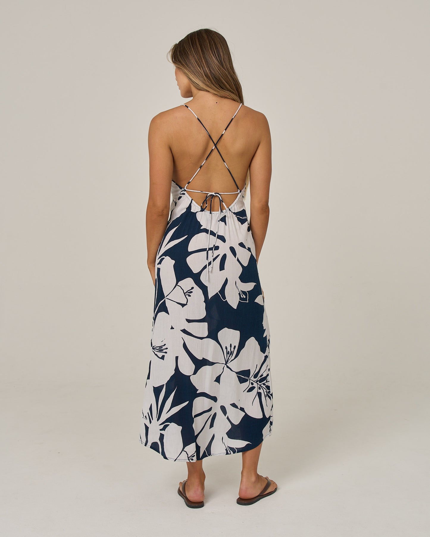 A woman stands facing away, wearing the Salty Crew Graphic Bloom Dress in Faded Mauve, which features cross-back straps and a floral design. She has straight, long hair and pairs the dress with black flip-flops, standing against a plain light background.
