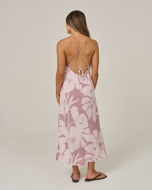 A woman stands with her back to the camera, wearing the Salty Crew Graphic Bloom Dress in dark navy, featuring cross-back straps, paired with black sandals against a plain background.