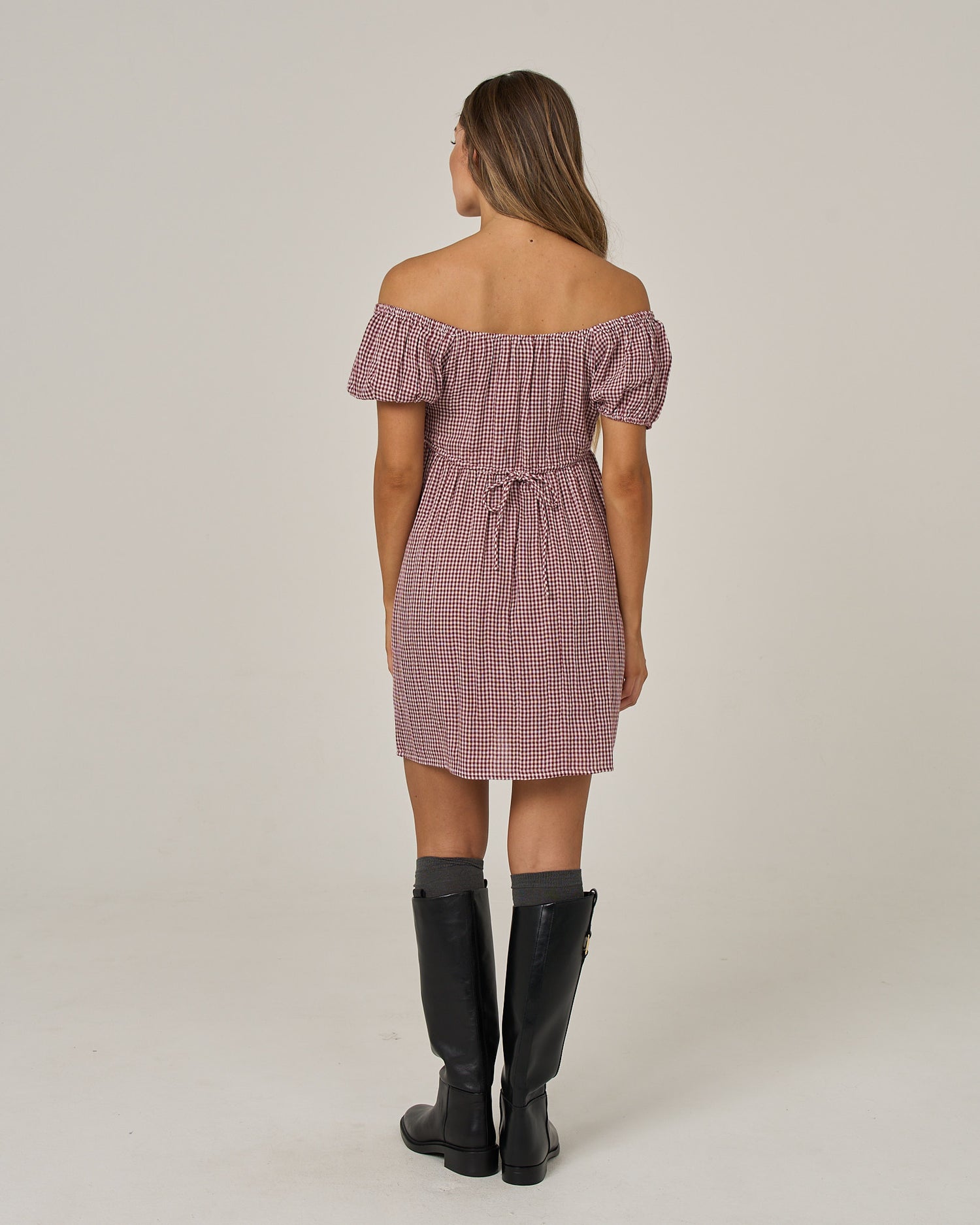 A woman with long hair is seen from behind, wearing the Salty Crew Fresh Catch Mini Dress in maroon—an off-the-shoulder, short baby doll style featuring an adjustable tie back—paired with black knee-high boots, posed against a plain background.