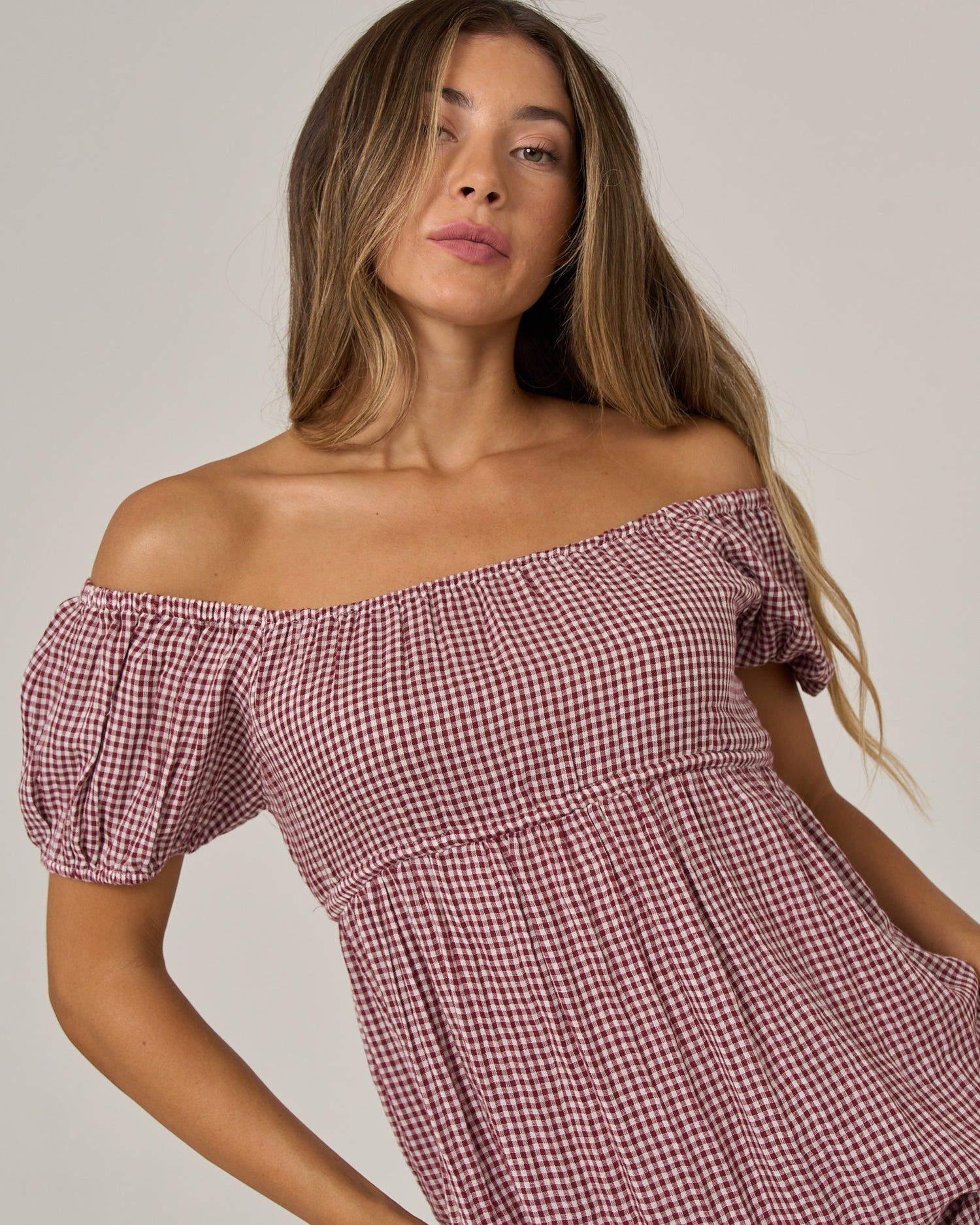 A woman with long brown hair wears the Salty Crew Fresh Catch Mini Dress in maroon, an off-the-shoulder, short-sleeved baby doll style featuring an adjustable tie back and a red and white gingham pattern, as she poses against a plain background.