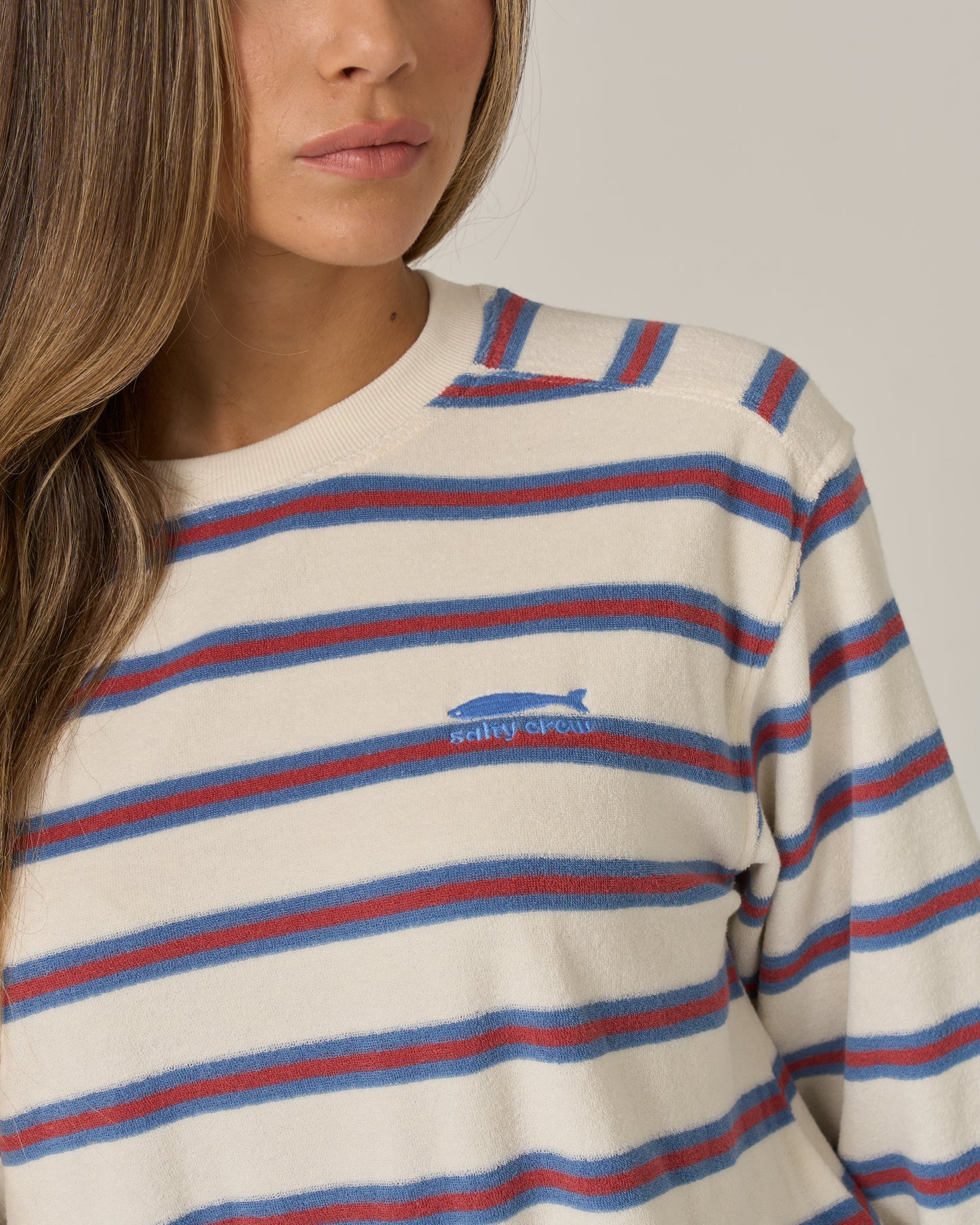 A woman with long brown hair wears the Salty Crew Night N Day Crew in Off White, featuring red and blue terry stripes and a small blue fish logo with "Salty Crew" text on the chest. The background is plain and neutral.