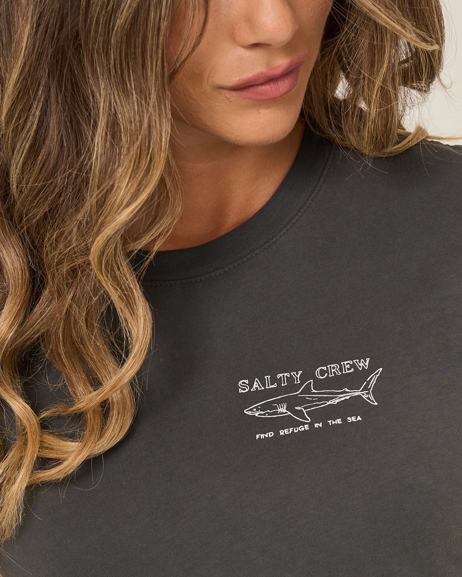 A woman with wavy, light brown hair wears the Salty Crew Shark Anatomy Modern Tee in charcoal, a fitted cotton jersey with cap sleeves featuring a white shark line drawing and "Find Refuge in the Sea.