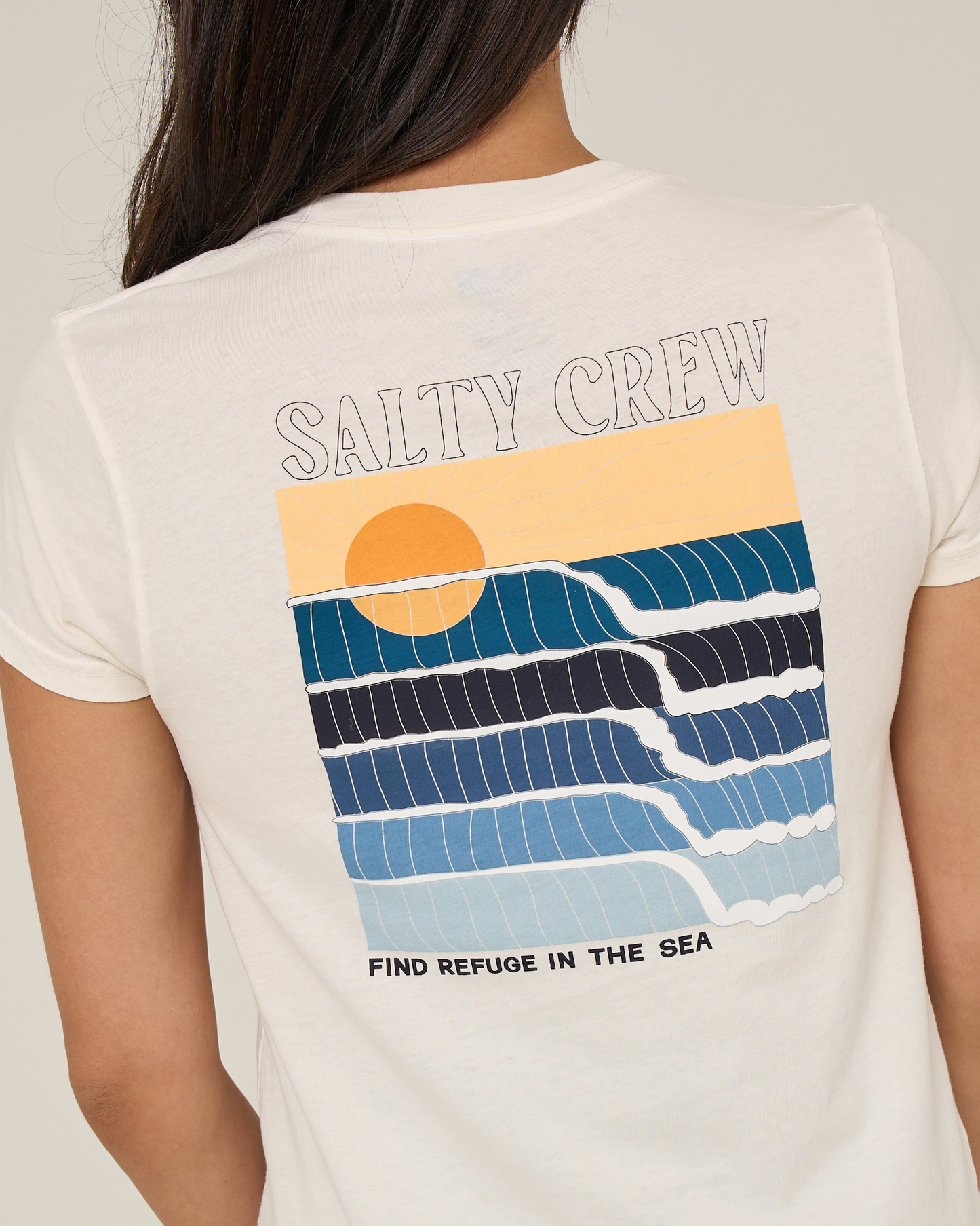 Someone wearing the Salty Crew Retro Wave Modern Tee in off white, featuring a screen print of ocean waves, a sun, and the text SALTY CREW and FIND REFUGE IN THE SEA on the back.
