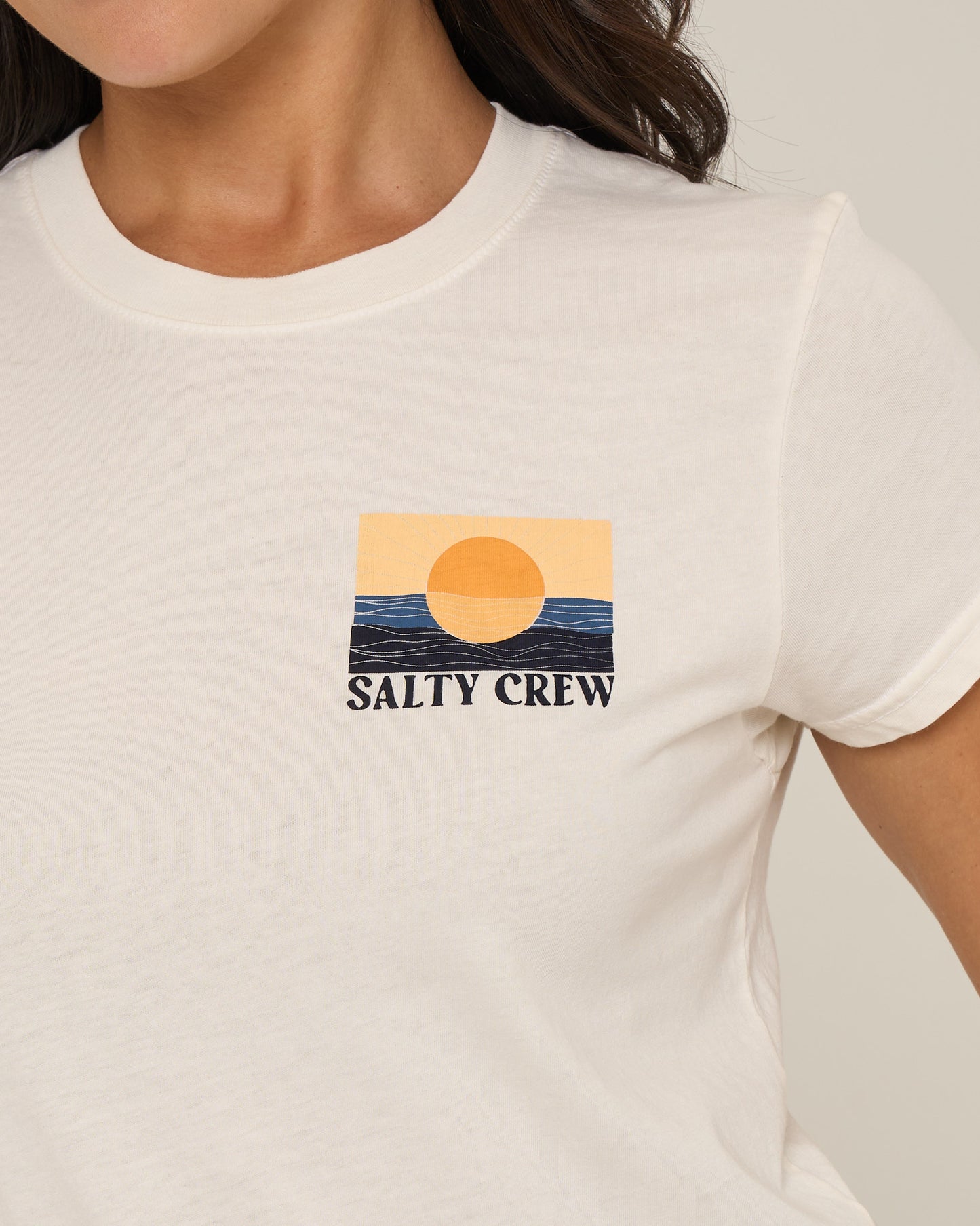 Someone is wearing the Salty Crew Retro Wave Modern Tee in off-white, featuring a stylized sunset over waves and SALTY CREW screen printed on the chest.