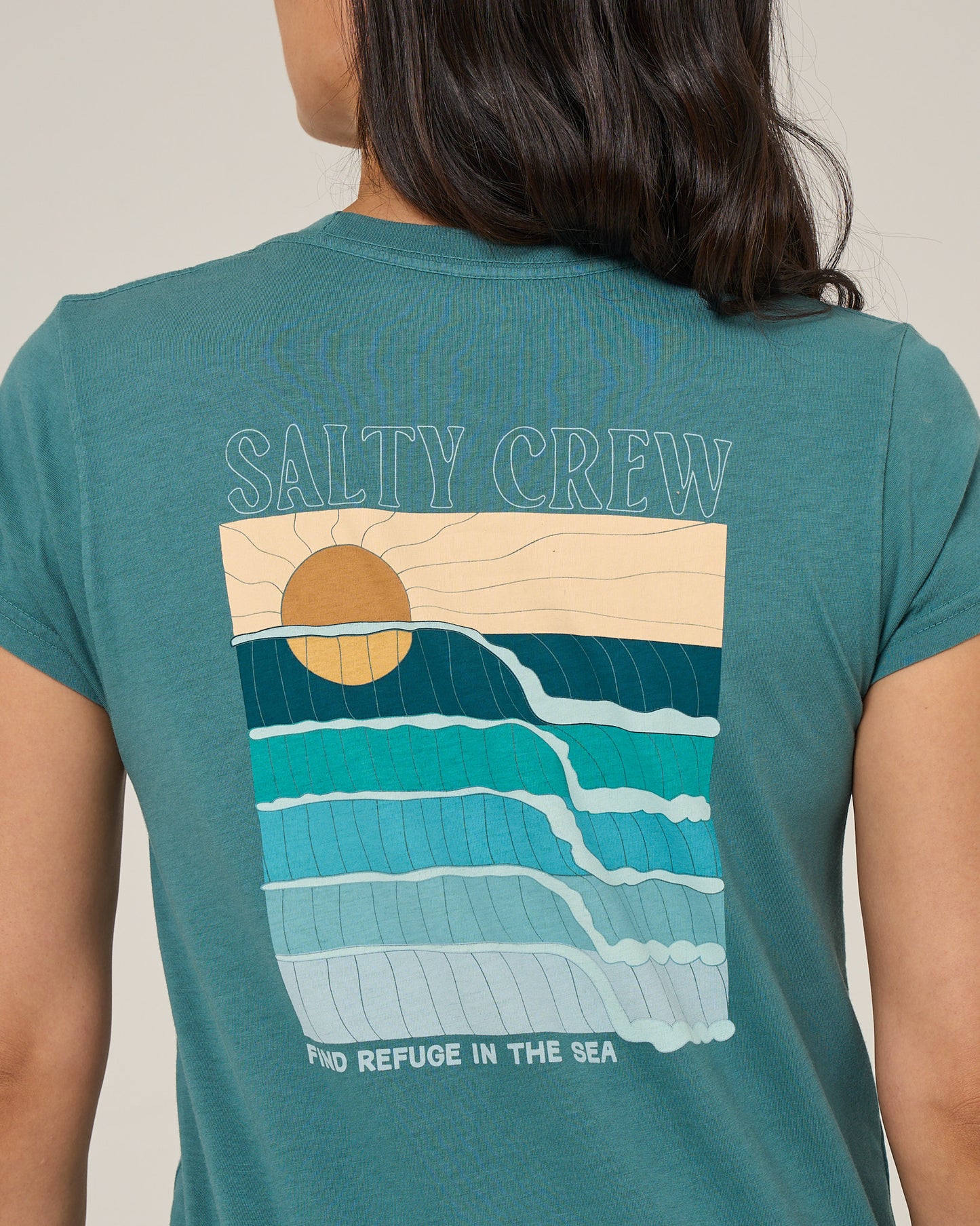 Someone is wearing the Salty Crew Retro Wave Modern Tee in teal, a cotton jersey with cap sleeves, featuring a sun over stylized waves. “SALTY CREW” appears above the print and “FIND REFUGE IN THE SEA” is written below.