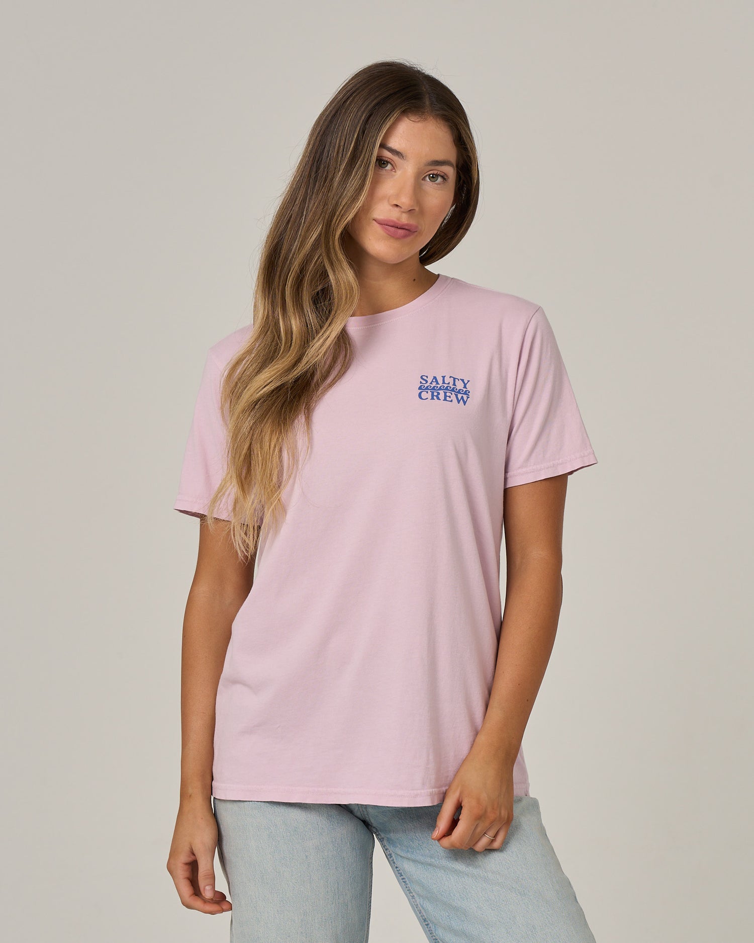 A woman with long, wavy hair wearing the Salty Crew Lobster Roll Boyfriend Tee in Pale Orchid and light blue jeans stands against a plain light gray background.