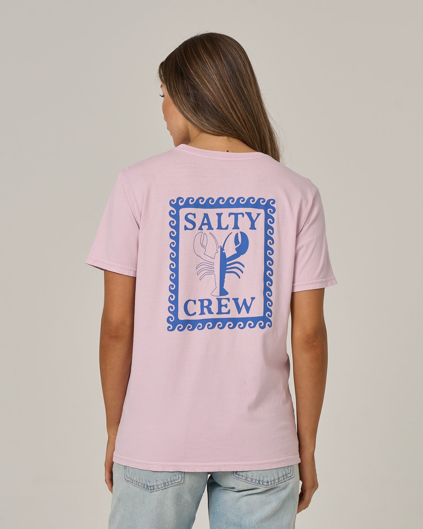 A person with long hair wears the Salty Crew Lobster Roll Boyfriend Tee in Pale Orchid, a light pink, short sleeve cotton jersey featuring a relaxed fit and a blue and white lobster graphic with Salty Crew branding on the back for casual comfort.
