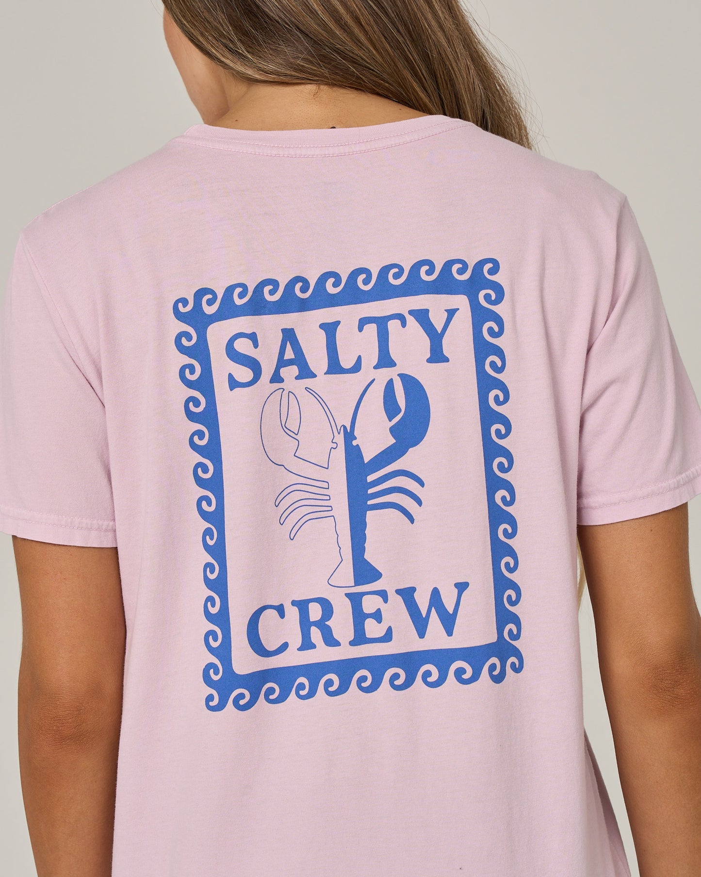 A person wearing the Salty Crew Lobster Roll Boyfriend Tee in Pale Orchid—a relaxed fit, pink cotton jersey shirt with short sleeves, featuring a blue lobster graphic and “Salty Crew” framed by a wave pattern on the back.