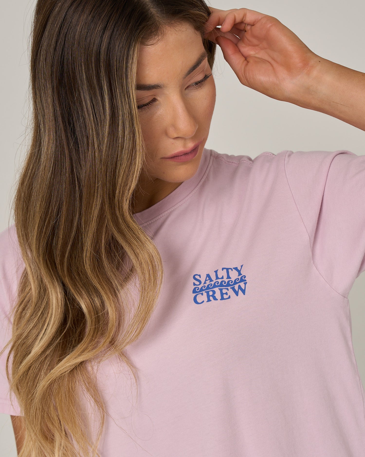 A woman with long, wavy hair in a relaxed fit, pale orchid Lobster Roll Boyfriend Tee by Salty Crew looks down and touches her head with one hand.