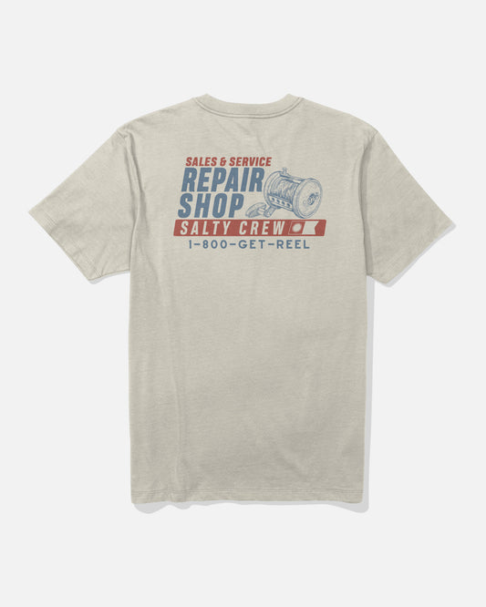 The Reel Repair Crew Tee - Bone by Salty Crew is a beige t-shirt crafted from soft, combed ringspun cotton for lasting comfort. Featuring “Repair Shop,” “Salty Crew,” and “1-800-GET-REEL” alongside a screenprinted illustration of a fishing reel on the back in blue and red text.