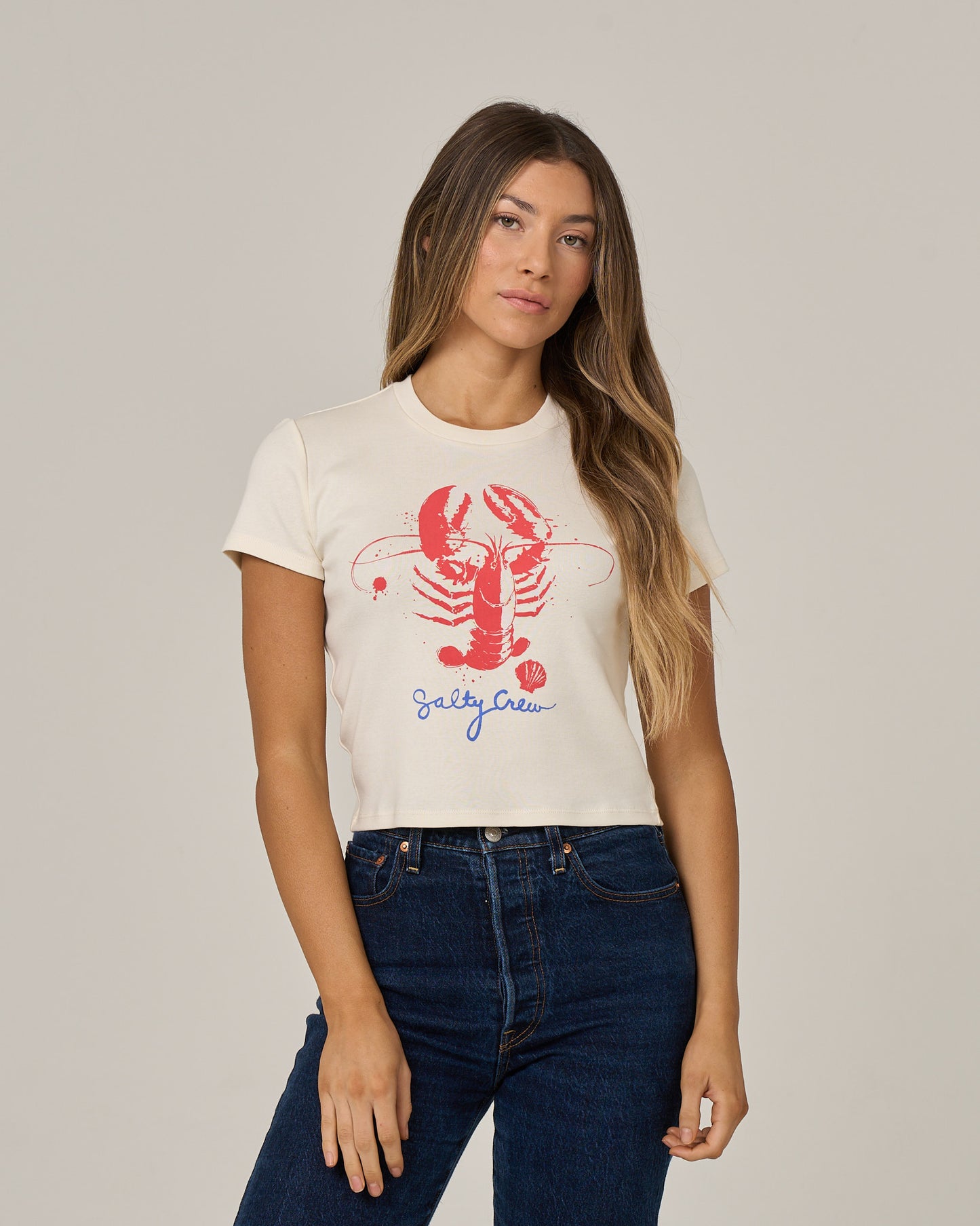 A woman with long brown hair poses in the Salty Crew Captains Special Baby Tee in Off White, a fitted short sleeve crop top featuring a vibrant red screen print of a lobster and seashell with “Salty Crew” written below in blue script. She pairs it with dark blue jeans against a light grey background.