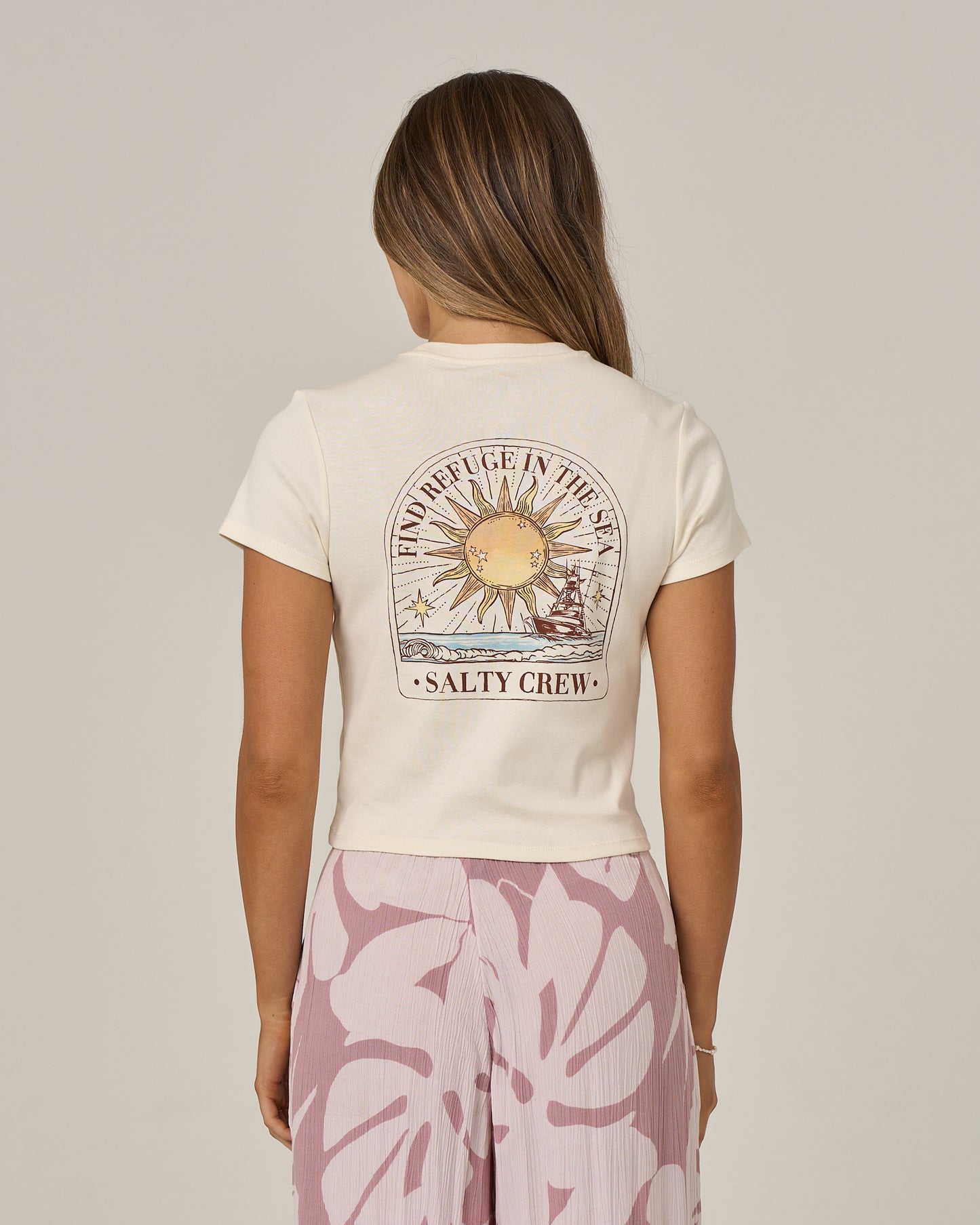 A woman with long hair wears the Salty Crew Celestial Sun Baby Tee in Off White, featuring a sun graphic and screen-printed “Find Refuge in the Sea” text. She also wears a pink skirt with a large floral pattern and is facing away from the camera.