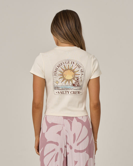 A woman with long hair wears the Salty Crew Celestial Sun Baby Tee in Off White, featuring a sun graphic and screen-printed “Find Refuge in the Sea” text. She also wears a pink skirt with a large floral pattern and is facing away from the camera.