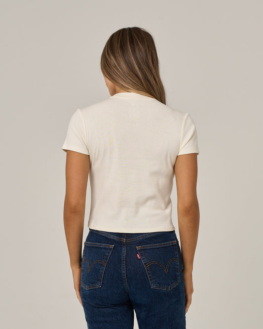 A person with long hair is standing facing away from the camera against a plain background, wearing blue jeans and the Salty Crew Captains Special Baby Tee in off white.