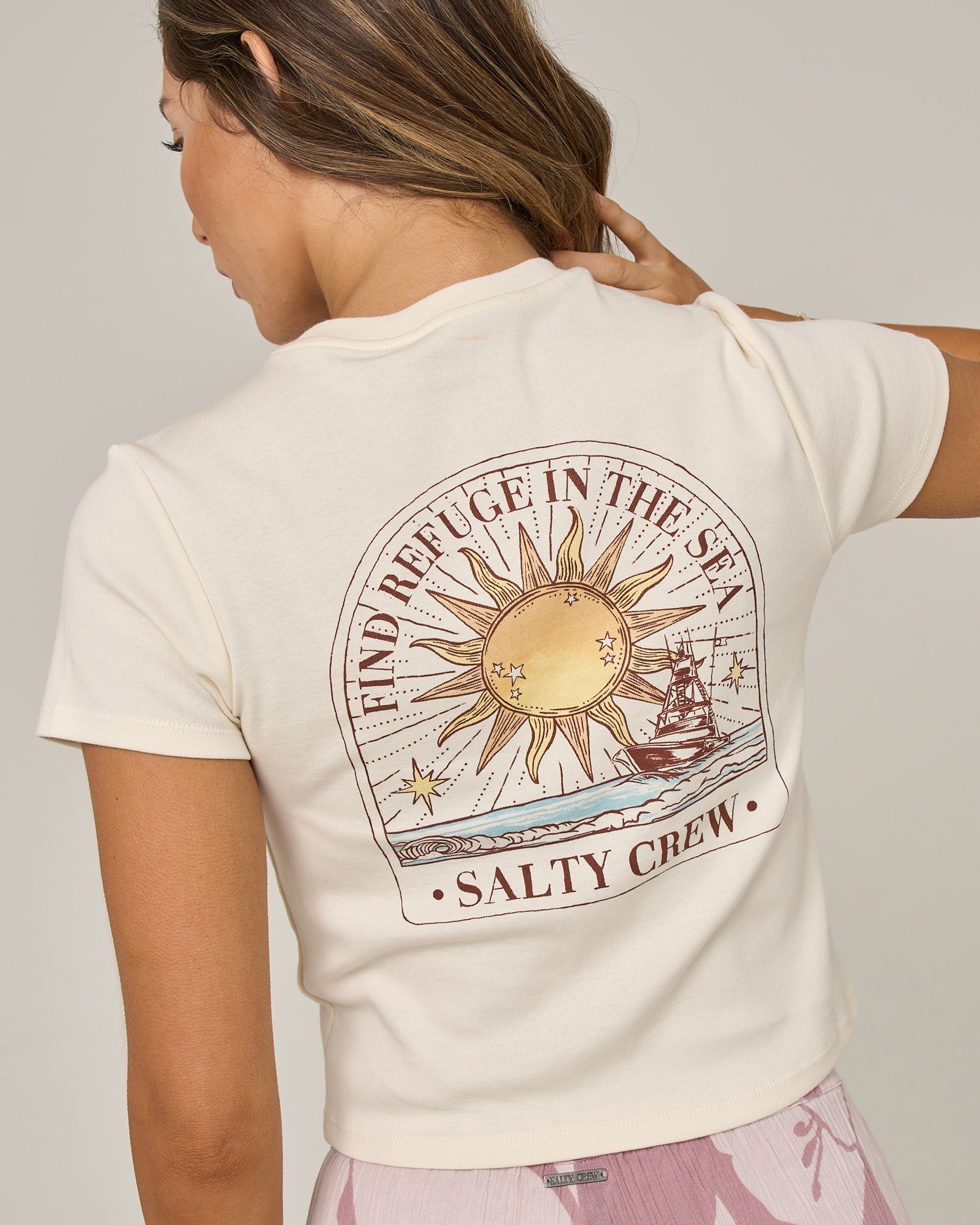 A woman faces away, wearing the Salty Crew Celestial Sun Baby Tee in Off White—a fitted cotton top featuring a sun, ship, and waves graphic with “FIND REFUGE IN THE SEA SALTY CREW” screen printed on the back. She touches her shoulder and pairs it with pink patterned pants.