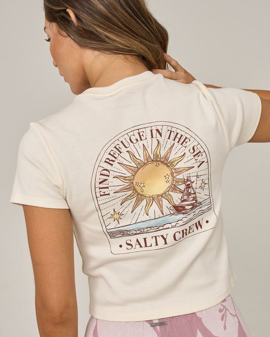 A woman faces away, wearing the Salty Crew Celestial Sun Baby Tee in Off White—a fitted cotton top featuring a sun, ship, and waves graphic with “FIND REFUGE IN THE SEA SALTY CREW” screen printed on the back. She touches her shoulder and pairs it with pink patterned pants.