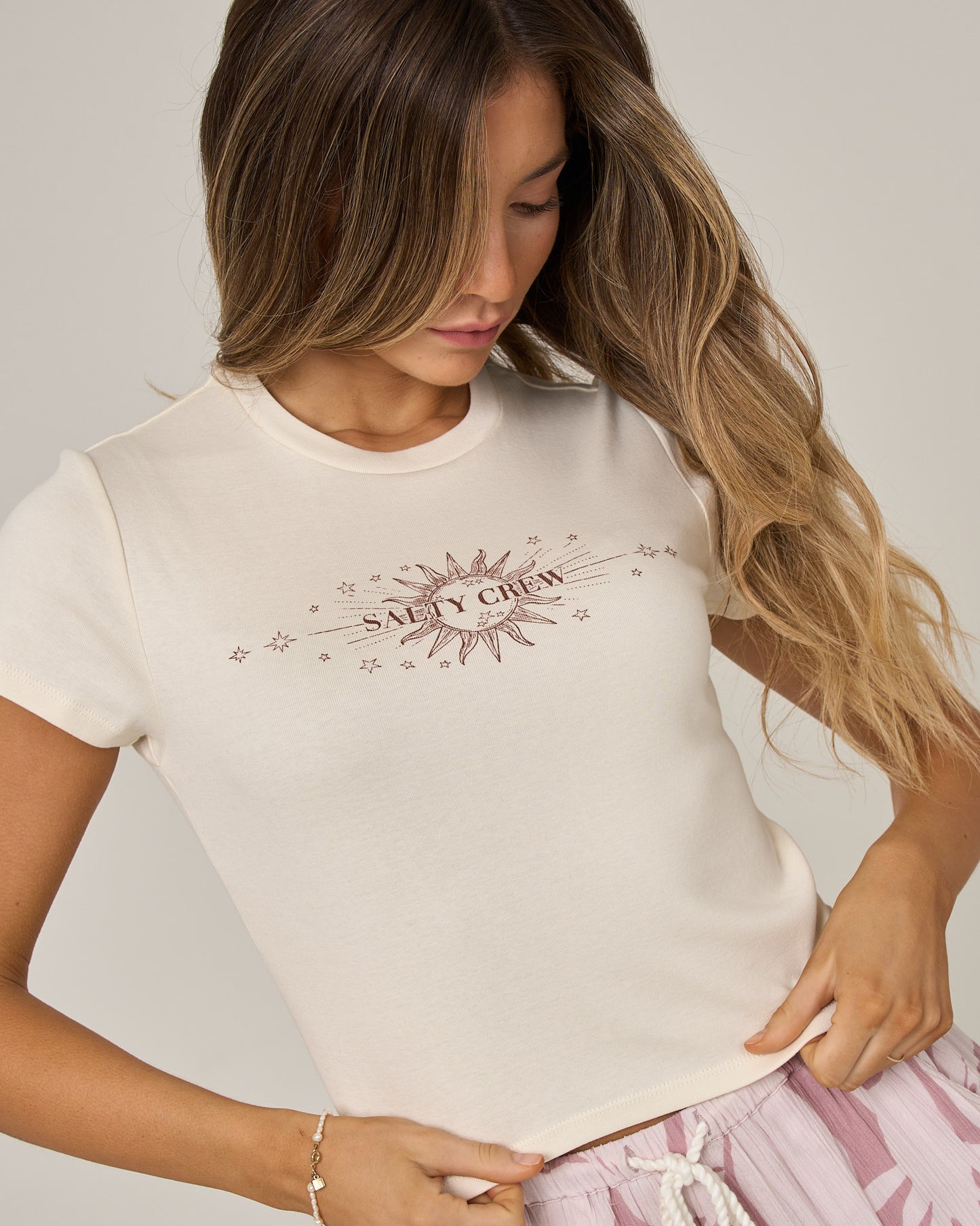 A woman with long, wavy hair looks down while wearing the Salty Crew Celestial Sun Baby Tee in Off White—a fitted, short-sleeve cotton top featuring a vibrant sunburst graphic and “Salty Crew” screen print—paired with a light pink drawstring skirt.
