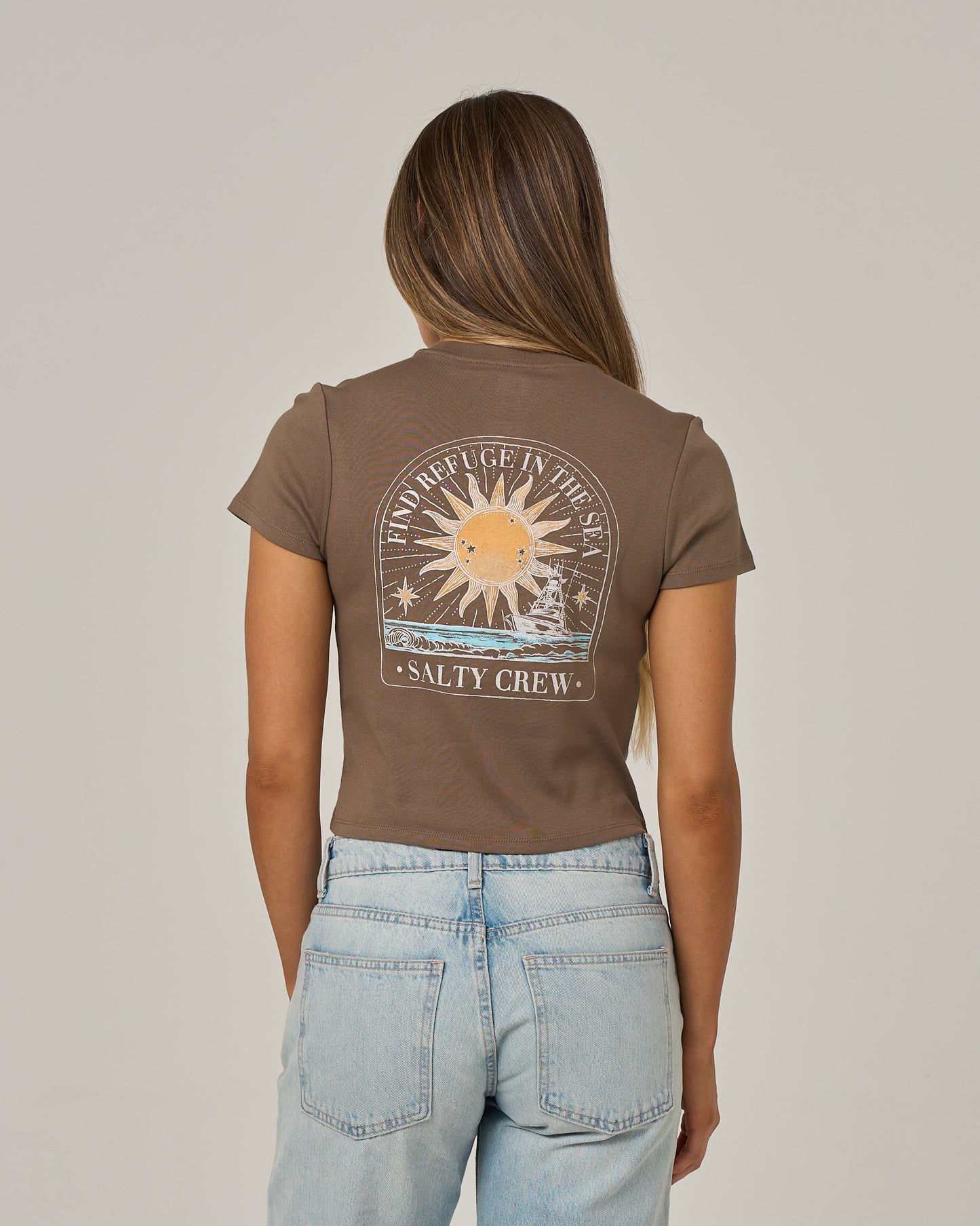 A woman with long, straight hair wears the Salty Crew Celestial Sun Baby Tee in washed brown, featuring a sun and wave graphic with SALTY CREW text on the back. She pairs it with light blue jeans and stands against a plain background.