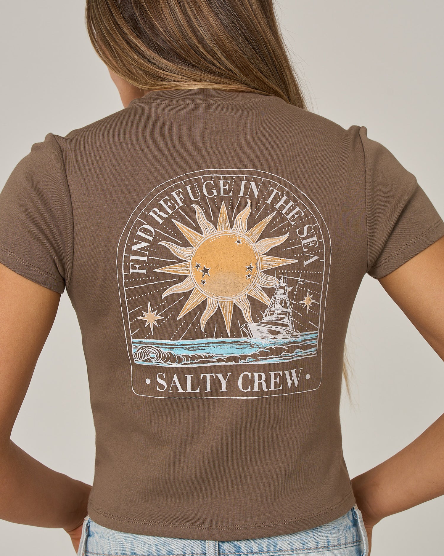 A woman is seen from the back wearing the Salty Crew Celestial Sun Baby Tee in washed brown. This fitted, short sleeve cotton top features a vibrant screen print on the back depicting a sun, ocean waves, and a ship, along with the phrases “FIND REFUGE IN THE SEA” and “SALTY CREW.”.