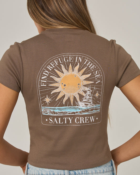 A woman is seen from the back wearing the Salty Crew Celestial Sun Baby Tee in washed brown. This fitted, short sleeve cotton top features a vibrant screen print on the back depicting a sun, ocean waves, and a ship, along with the phrases “FIND REFUGE IN THE SEA” and “SALTY CREW.”.