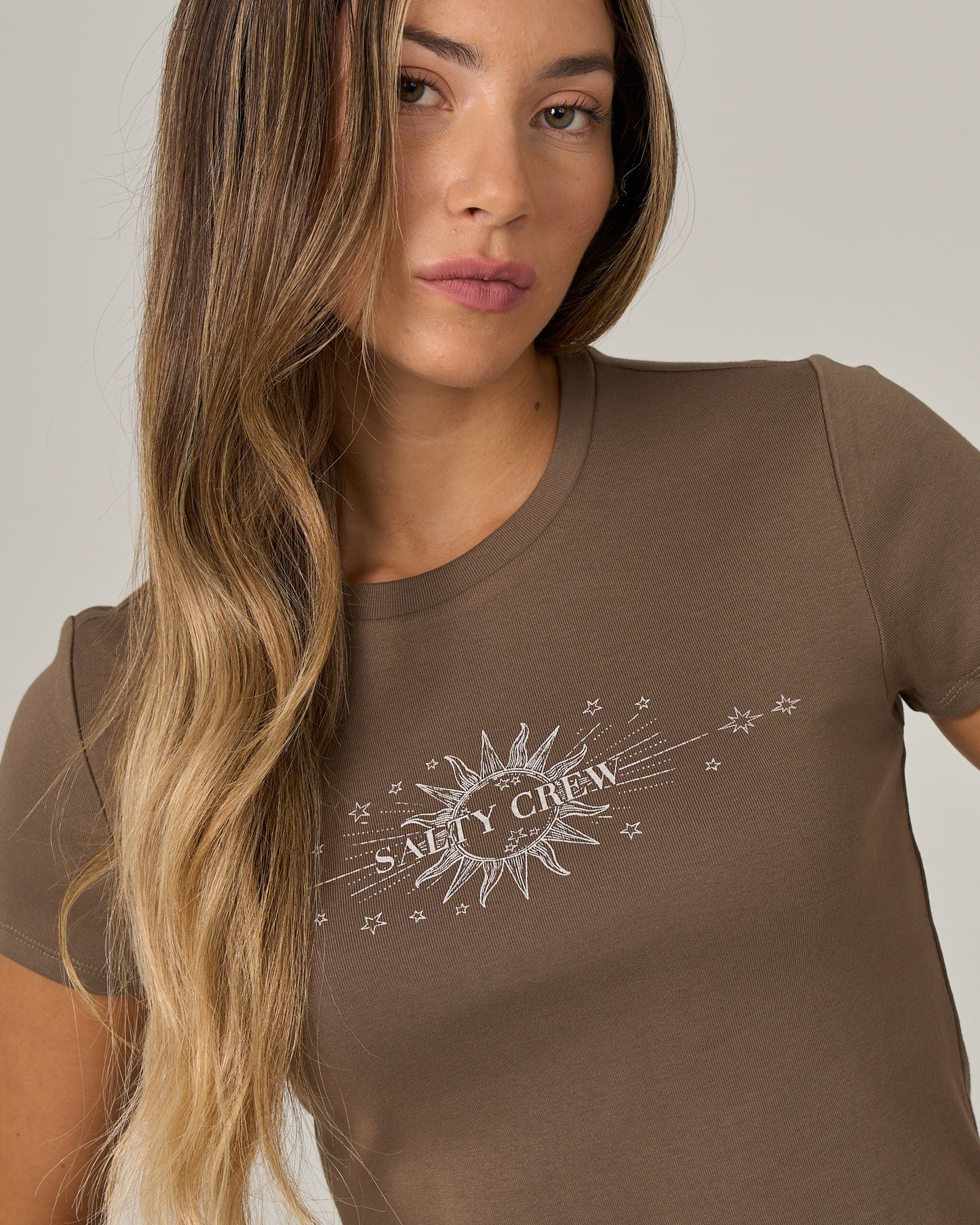A woman with long, wavy hair wears the Salty Crew Celestial Sun Baby Tee in washed brown—a fitted, short-sleeve cotton tee featuring a sun and stars screen print graphic and the Salty Crew logo on the front.