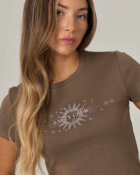 A woman with long, wavy hair wears the Salty Crew Celestial Sun Baby Tee in washed brown—a fitted, short-sleeve cotton tee featuring a sun and stars screen print graphic and the Salty Crew logo on the front.