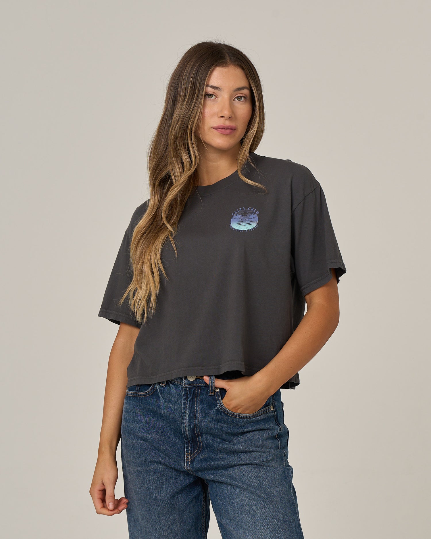 A woman with long, wavy hair wears the Salty Crew Tropics Skimmer Tee in charcoal, which features a small blue graphic, paired with blue jeans as she stands against a plain, light background.