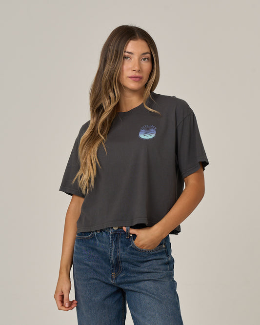 A woman with long, wavy hair wears the Salty Crew Tropics Skimmer Tee in charcoal, which features a small blue graphic, paired with blue jeans as she stands against a plain, light background.