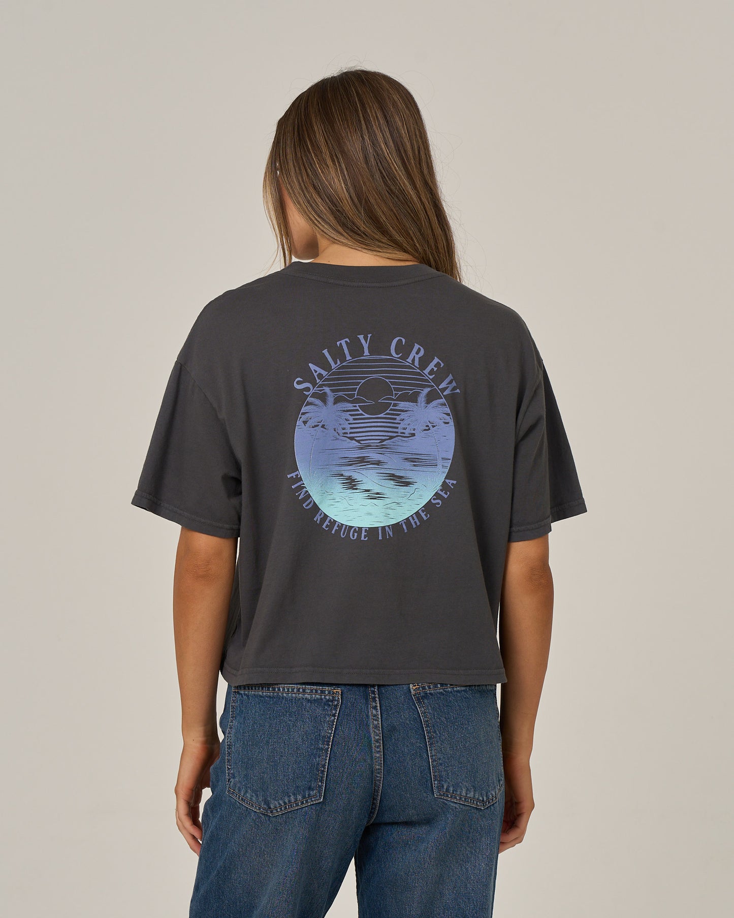 A person is shown from behind against a plain background, wearing the Salty Crew Tropics Skimmer Tee in Charcoal featuring a circular ocean graphic and text, paired with blue jeans.