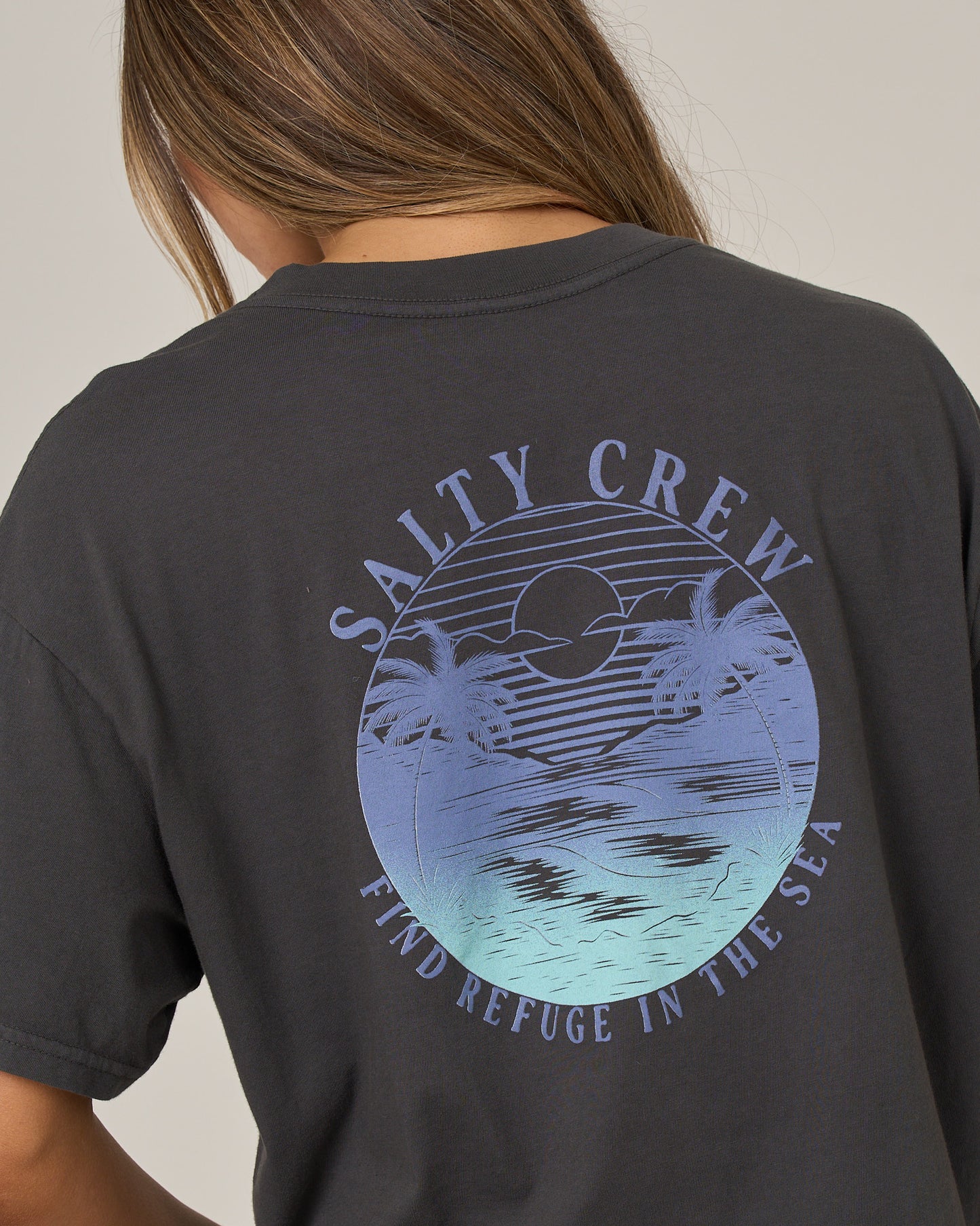 A person wears the Salty Crew Tropics Skimmer Tee in charcoal—a dark, boxy-fit t-shirt featuring a blue graphic of a sunset, ocean, and palm trees. The screen-printed design displays “SALTY CREW” above and “FIND REFUGE IN THE SEA” below the image.