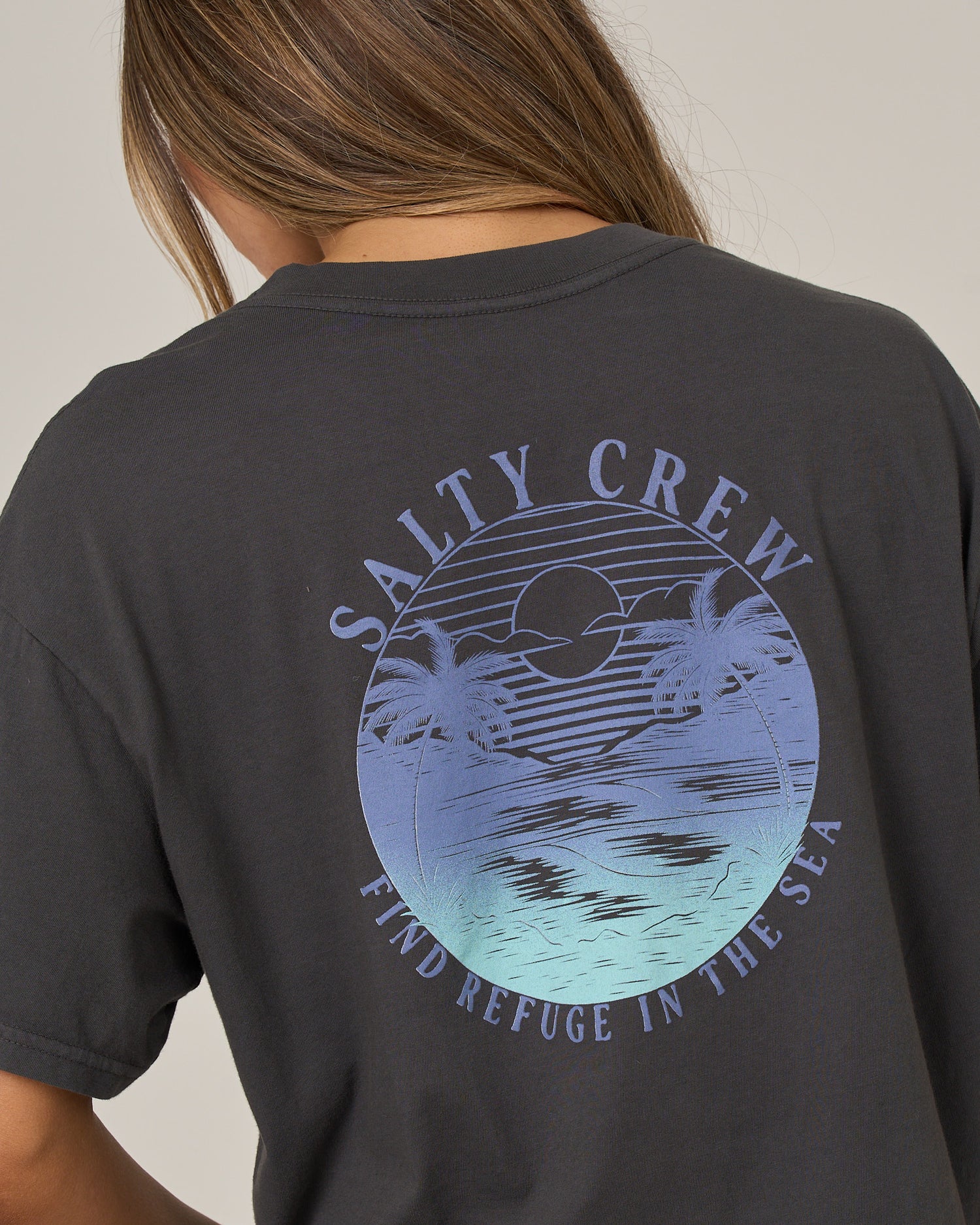 A person wears the Salty Crew Tropics Skimmer Tee in charcoal—a dark, boxy-fit t-shirt featuring a blue graphic of a sunset, ocean, and palm trees. The screen-printed design displays “SALTY CREW” above and “FIND REFUGE IN THE SEA” below the image.
