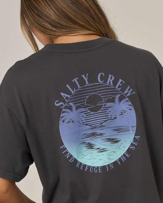 A person wears the Salty Crew Tropics Skimmer Tee in charcoal—a dark, boxy-fit t-shirt featuring a blue graphic of a sunset, ocean, and palm trees. The screen-printed design displays “SALTY CREW” above and “FIND REFUGE IN THE SEA” below the image.