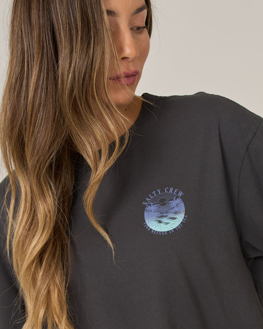 A woman with long, wavy hair looks down, wearing the Salty Crew Tropics Skimmer Tee in charcoal, featuring a wave and sun graphic screen printed on the chest.