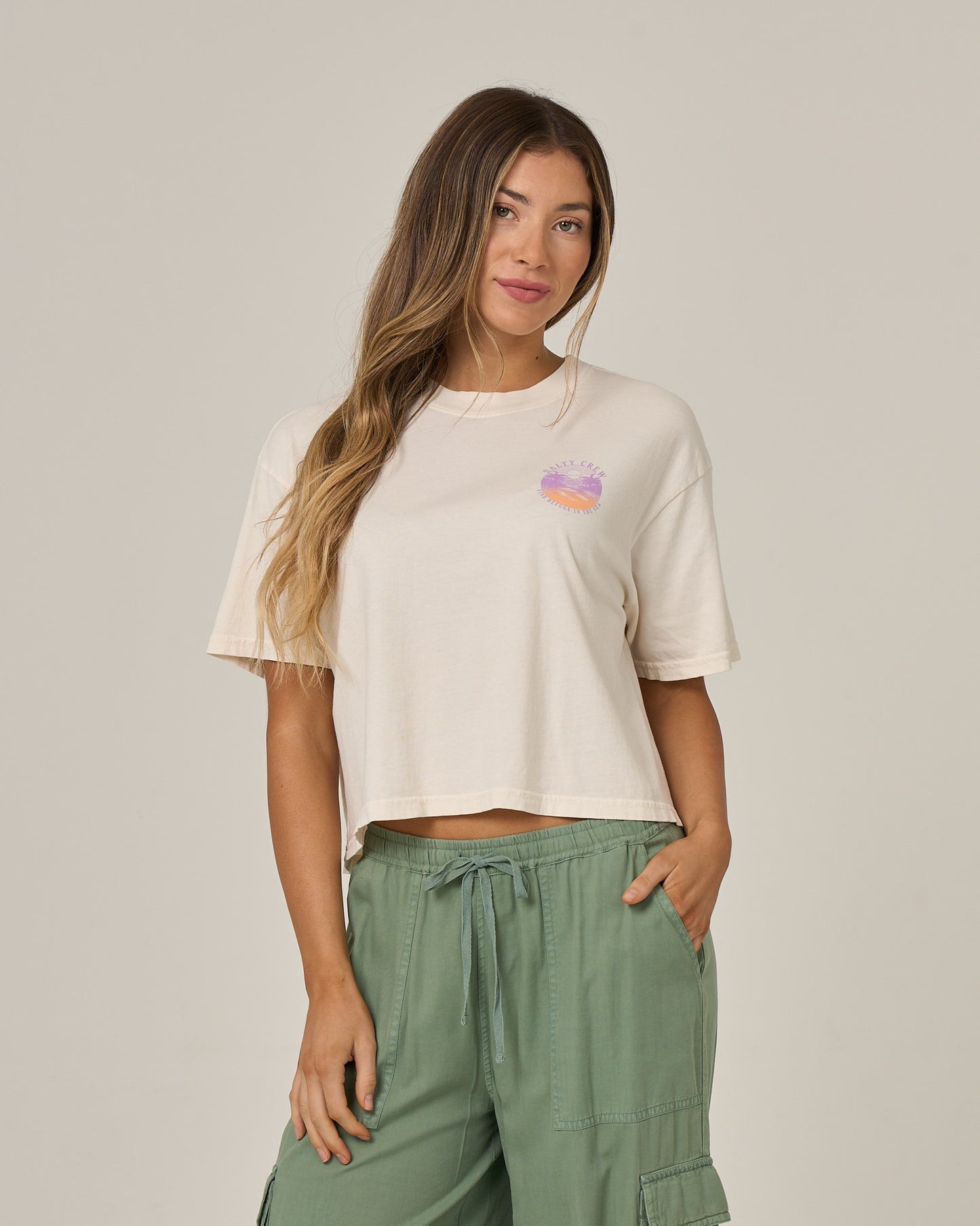A woman with long brown hair wears the Salty Crew Tropics Skimmer Tee in Off White, featuring a small, colorful graphic on the chest, paired with green drawstring cargo pants as she stands against a plain light background.