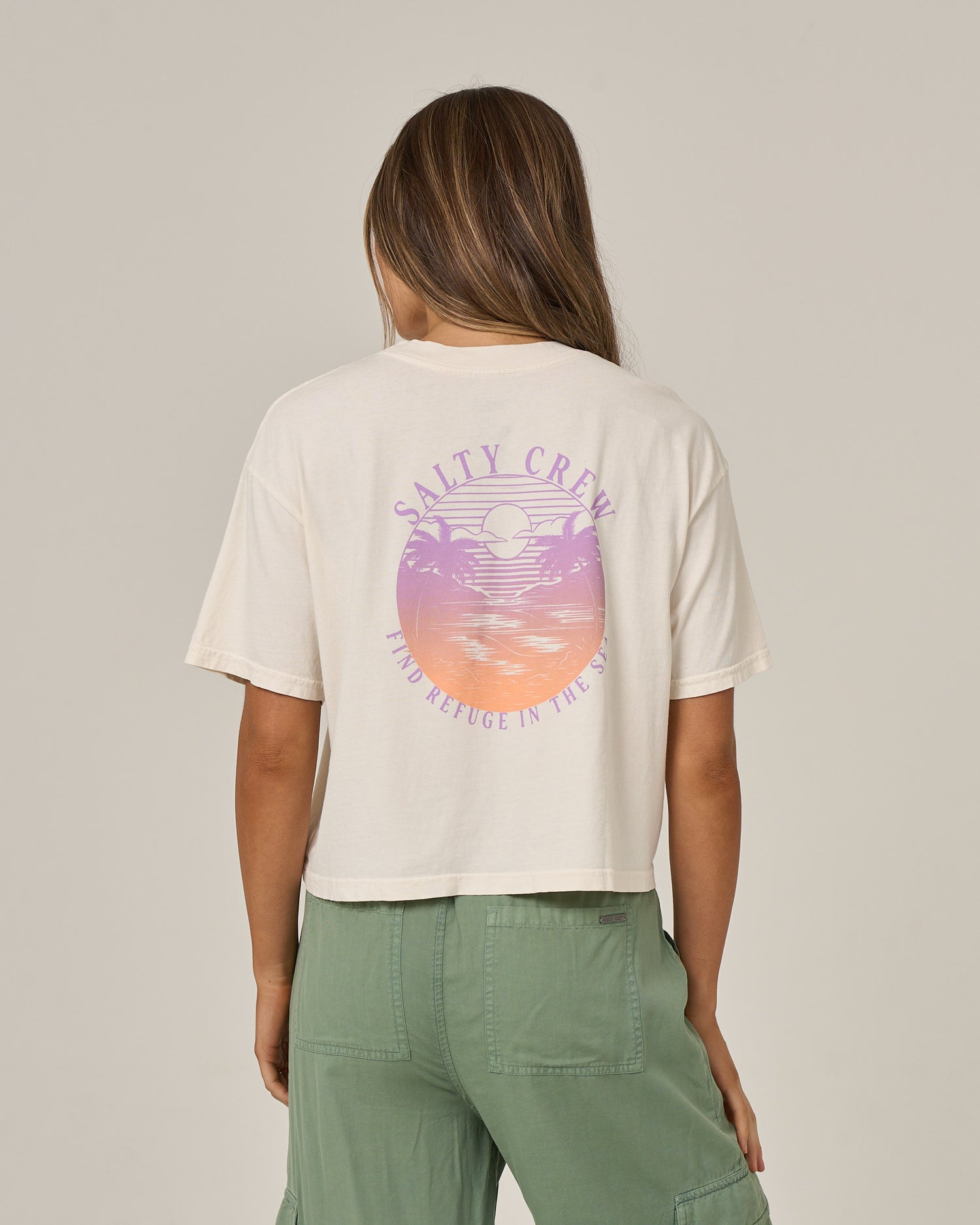 A person with long hair wears the Salty Crew Tropics Skimmer Tee in Off White, a cotton jersey shirt featuring a pastel sunset and ocean graphic with “Find Refuge in the Sea” on the back, paired with green pants.