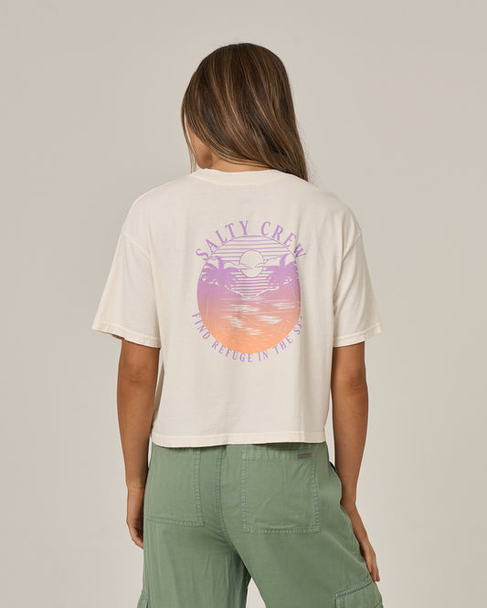 A person with long hair wears the Salty Crew Tropics Skimmer Tee in Off White, a cotton jersey shirt featuring a pastel sunset and ocean graphic with “Find Refuge in the Sea” on the back, paired with green pants.
