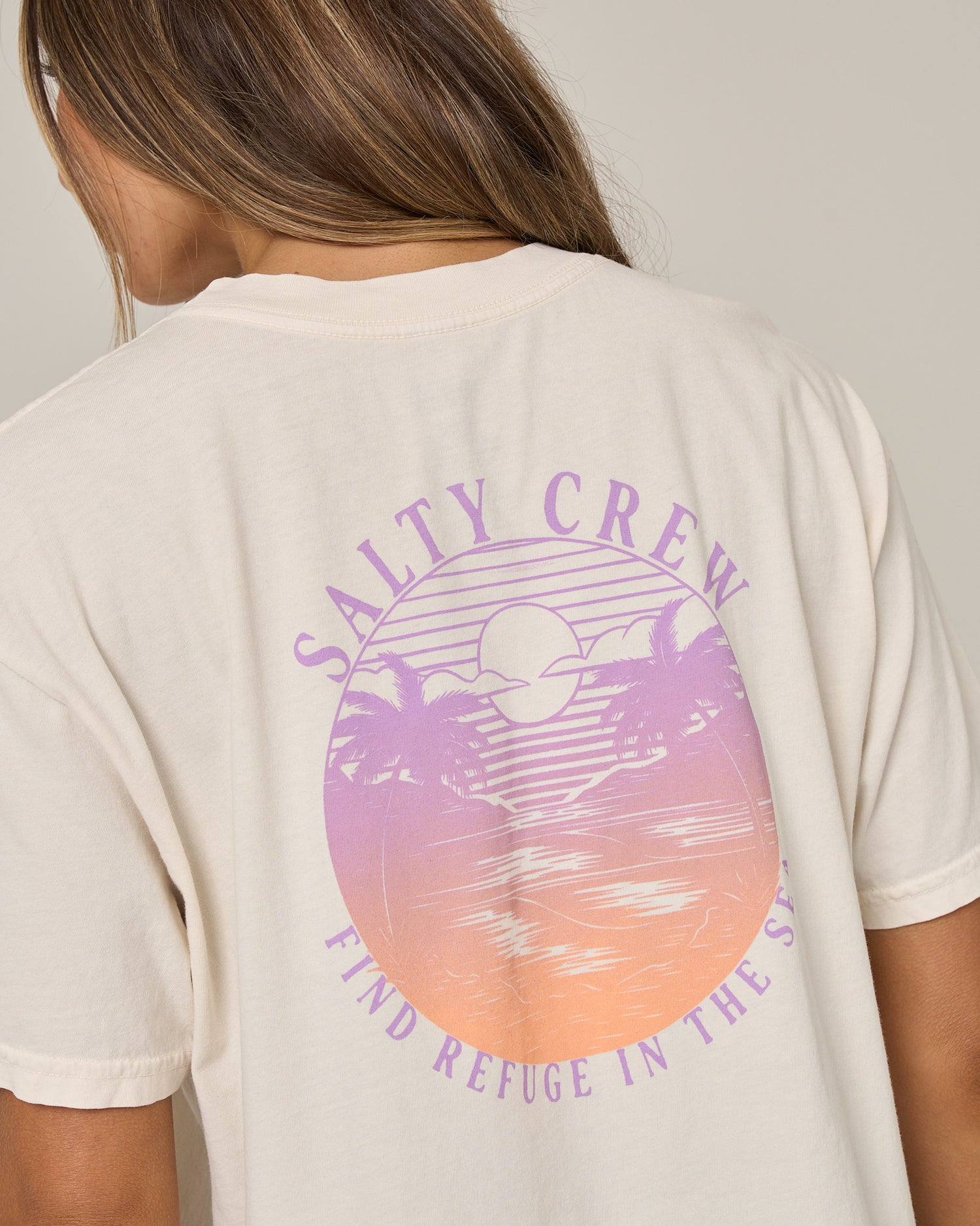 A person wearing the Salty Crew Tropics Skimmer Tee in off white, a cream cotton jersey shirt featuring a screen print graphic of a sunset, palm trees, and wavy lines, with “SALTY CREW” printed above and “FIND REFUGE IN THE SEA” below the image.