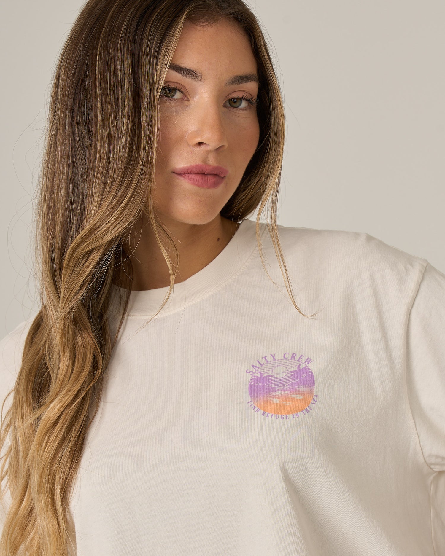 A woman with long, wavy hair wears the Salty Crew Tropics Skimmer Tee in Off White—a cotton jersey top featuring a small, pastel sunset and wave graphic on the chest—while standing against a plain, light background.