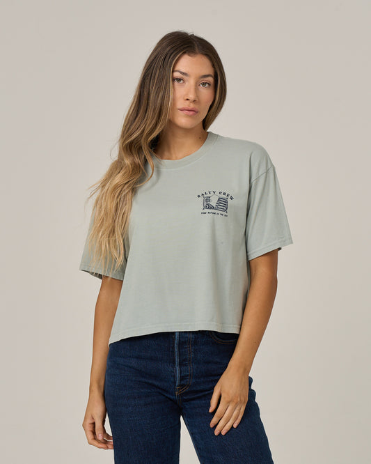 A woman with long wavy hair wears the Salty Crew Cute Tin Skimmer Tee in Aqua Grey—a light green, boxy-fitting tee made from soft cotton jersey with a small screen print graphic and text on the left chest—paired with dark blue jeans, standing against a plain light background.