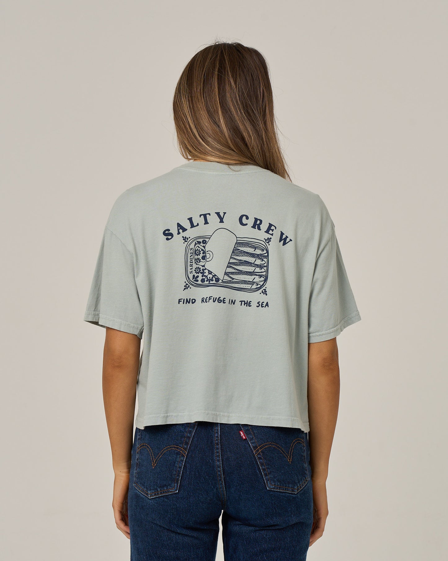 A woman stands facing away, wearing the Salty Crew Cute Tin Skimmer Tee in aqua grey—a boxy top featuring “Salty Crew” and “Find Refuge in the Sea” text with a screen-printed fish graphic on the back—paired with high-waisted blue jeans.