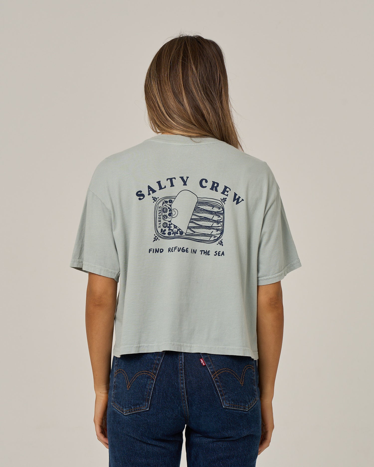 A woman stands facing away, wearing the Salty Crew Cute Tin Skimmer Tee in aqua grey—a boxy top featuring “Salty Crew” and “Find Refuge in the Sea” text with a screen-printed fish graphic on the back—paired with high-waisted blue jeans.
