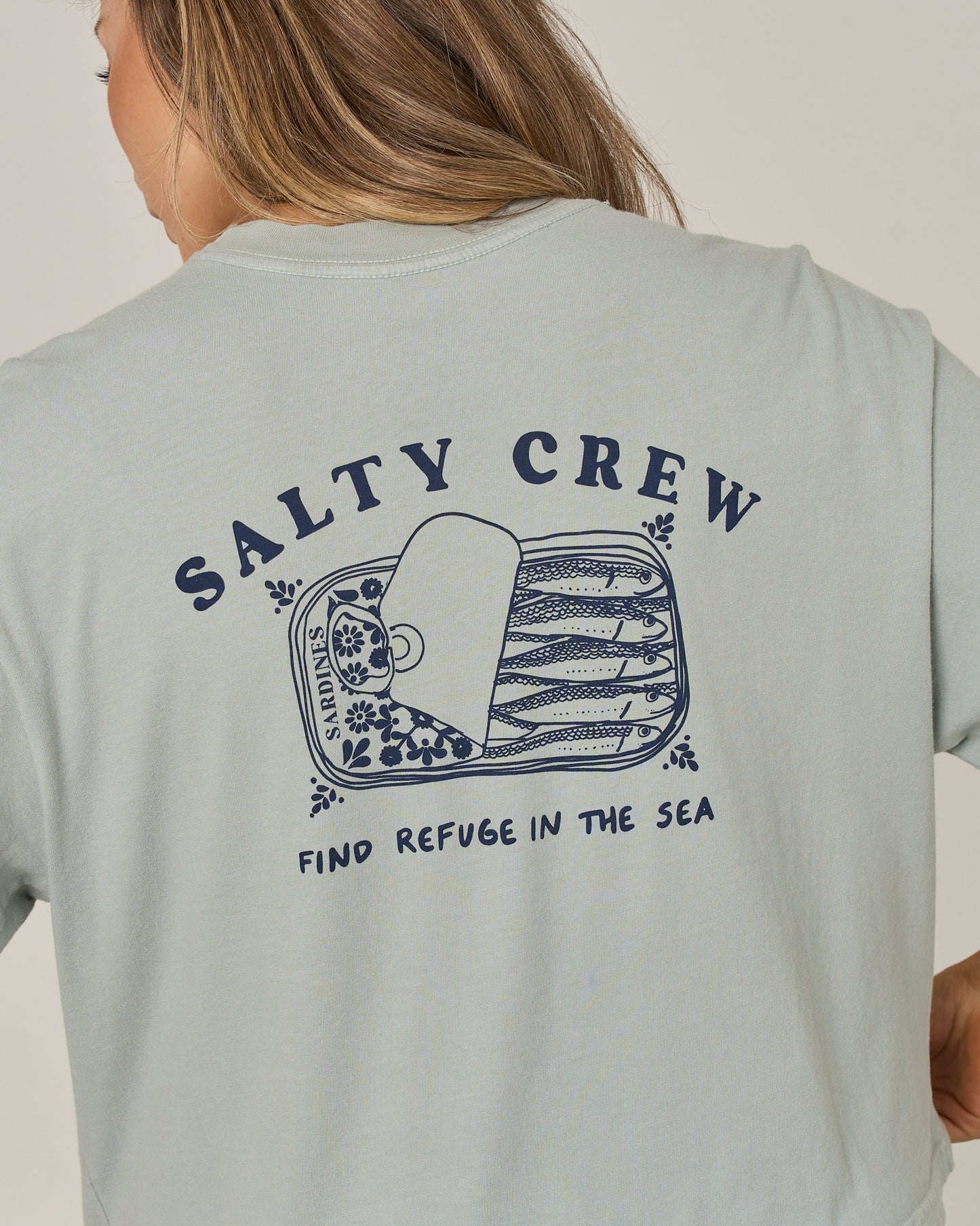 A person wearing the Cute Tin Skimmer Tee in aqua grey by Salty Crew, featuring a sardine can illustration and the text “Salty Crew” and “Find Refuge in the Sea” screen printed on the back.