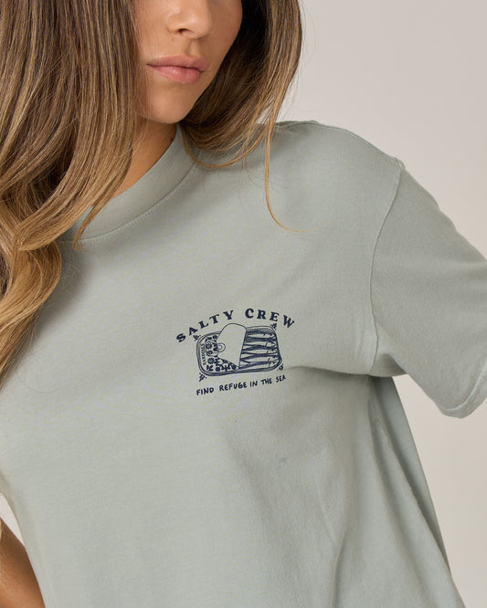 A woman wearing the Salty Crew Cute Tin Skimmer Tee in Aqua Grey—a cotton jersey shirt featuring a small fish graphic and “Find Refuge in the Sea” text on the left chest. The image is cropped at the shoulders.