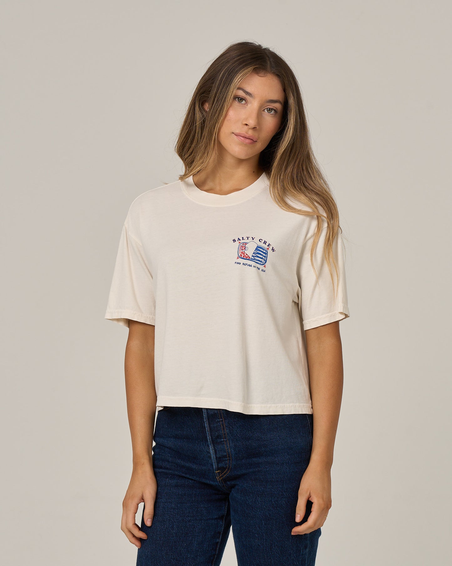 A woman stands against a plain light background, looking at the camera with a neutral expression, wearing the Salty Crew Cute Tin Skimmer Tee in Off White—a boxy style featuring a small screen print graphic and text on the left chest.